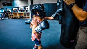 Young girl wearing boxing headgear and gloves punches a hanging bag while a coach holds it for guidance in a gymenario setting.