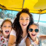 Woman and two kids smiling and taking a selfie under a bright yellow canopy on a seaside boardwalk with the beach in the background