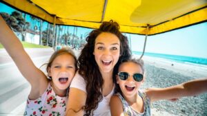 Woman and two kids smiling and taking a selfie under a bright yellow canopy on a seaside boardwalk with the beach in the background