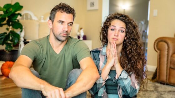 A man and a woman sit on the living-room floor looking distressed, with the woman’s hands pressed together in a pleading gesture.
