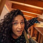 Woman with curly hair in a wooden attic, reaching forward with a surprised expression as she looks up and to the side.