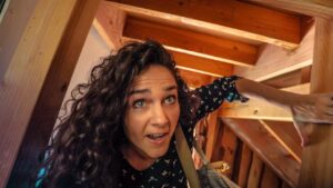 Woman with curly hair in a wooden attic, reaching forward with a surprised expression as she looks up and to the side.