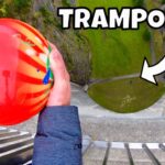 Person grips a bright red-orange ball while looking down from a tall structure toward a green circular area below with a 'TRAMPOLINE' overlay text.