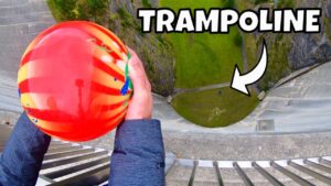 Person grips a bright red-orange ball while looking down from a tall structure toward a green circular area below with a 'TRAMPOLINE' overlay text.