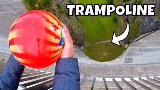 Person grips a bright red-orange ball while looking down from a tall structure toward a green circular area below with a 'TRAMPOLINE' overlay text.