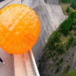 Man on a mountain road with a giant orange inflatable ball rolling toward him, arms raised.