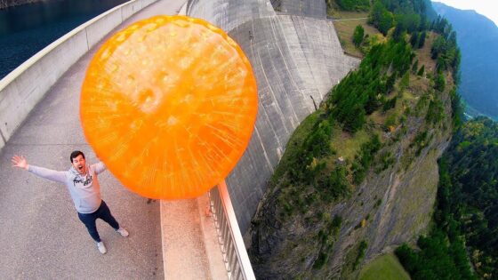 Man on a mountain road with a giant orange inflatable ball rolling toward him, arms raised.