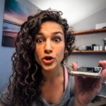 Woman with curly hair holding a smartphone toward the camera, speaking excitedly in a home office or bedroom setting.