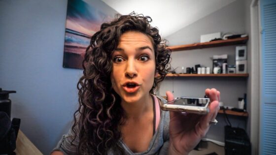 Woman with curly hair holding a smartphone toward the camera, speaking excitedly in a home office or bedroom setting.