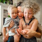 Mother holding two toddlers close as they examine something in her hands in a bright living room.