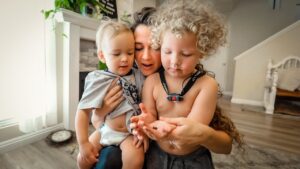Mother holding two toddlers close as they examine something in her hands in a bright living room.