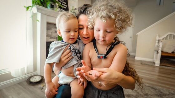 Mother holding two toddlers close as they examine something in her hands in a bright living room.