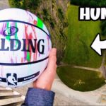 Hands hold a colorful Spalding basketball over a high drop, with a rocky cliff and path below in view.