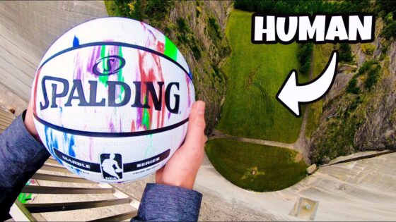Hands hold a colorful Spalding basketball over a high drop, with a rocky cliff and path below in view.