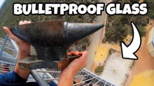 Person on a high balcony grips a heavy metal anvil with a view of ground far below; thumbnail reads 'BULLETPROOF GLASS'.
