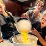 Three girls bake in a kitchen; one pours yellow batter into a bowl of flour while two others watch.