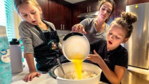 Three girls bake in a kitchen; one pours yellow batter into a bowl of flour while two others watch.