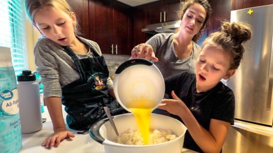 Three girls bake in a kitchen; one pours yellow batter into a bowl of flour while two others watch.