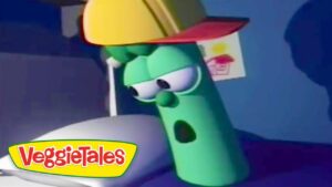Green cucumber character from VeggieTales wearing a yellow construction helmet, leaning on a white surface with the VeggieTales logo visible in the corner.