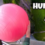Person gripping a large pink ball while hanging over the edge of a tall building, looking down at a grassy area far below.
