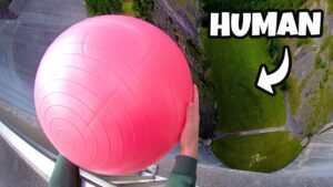 Person gripping a large pink ball while hanging over the edge of a tall building, looking down at a grassy area far below.