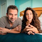A smiling couple lying on a bed, leaning on their elbows, in a bright turquoise bedroom.