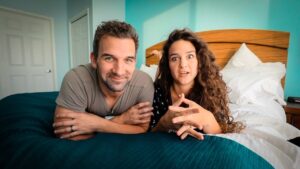 A smiling couple lying on a bed, leaning on their elbows, in a bright turquoise bedroom.