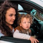 Mother with curly hair leaning out of a car window, watching something outside with a young child beside her in the passenger seat or back seat area of the car onlook.
