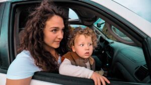Mother with curly hair leaning out of a car window, watching something outside with a young child beside her in the passenger seat or back seat area of the car onlook.