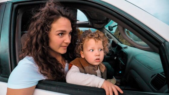 Mother with curly hair leaning out of a car window, watching something outside with a young child beside her in the passenger seat or back seat area of the car onlook.