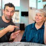 Man in a dark gray t‑shirt points at an older woman seated at a dining table; both smile at the camera in a cozy home setting.