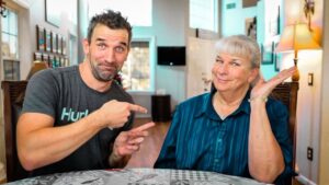 Man in a dark gray t‑shirt points at an older woman seated at a dining table; both smile at the camera in a cozy home setting.