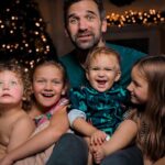 A father sits with four young children on a sofa, smiling, with a Christmas tree and twinkling lights in the background (holiday family photo).','A family portrait of a dad and his four kids cuddled together on a couch near warm Christmas lights.','Father with three daughters and a baby boy, all smiling, sitting close on a living room sofa by lit holiday decor.','A bundled-up family group on a couch: two older girls, a baby boy, a younger girl, with festive lights behind them.','Smiling father surrounded by his children on a couch, festive Christmas lights creating a warm holiday backdrop.