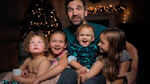A father sits with four young children on a sofa, smiling, with a Christmas tree and twinkling lights in the background (holiday family photo).','A family portrait of a dad and his four kids cuddled together on a couch near warm Christmas lights.','Father with three daughters and a baby boy, all smiling, sitting close on a living room sofa by lit holiday decor.','A bundled-up family group on a couch: two older girls, a baby boy, a younger girl, with festive lights behind them.','Smiling father surrounded by his children on a couch, festive Christmas lights creating a warm holiday backdrop.