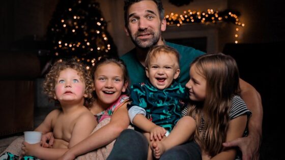 A father sits with four young children on a sofa, smiling, with a Christmas tree and twinkling lights in the background (holiday family photo).','A family portrait of a dad and his four kids cuddled together on a couch near warm Christmas lights.','Father with three daughters and a baby boy, all smiling, sitting close on a living room sofa by lit holiday decor.','A bundled-up family group on a couch: two older girls, a baby boy, a younger girl, with festive lights behind them.','Smiling father surrounded by his children on a couch, festive Christmas lights creating a warm holiday backdrop.