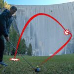 Man in a dark jacket tees off with a golf driver on a grassy field as a red trajectory arc shows the intended ball path toward a dam in the background, ending at a highlighted point on the slope.
