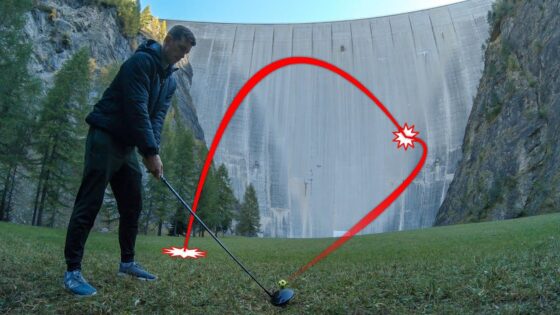 Man in a dark jacket tees off with a golf driver on a grassy field as a red trajectory arc shows the intended ball path toward a dam in the background, ending at a highlighted point on the slope.