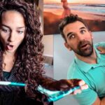 Woman with curly hair looks surprised while holding a turquoise item toward the camera; a man holds a smiling baby beside her in a kitchen/dining area.