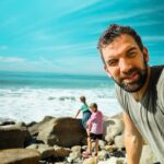 Man takes a selfie on a rocky beach while a pair of children play near the waves in the background.