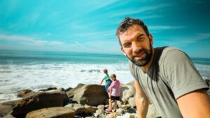 Man takes a selfie on a rocky beach while a pair of children play near the waves in the background.