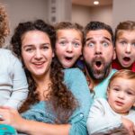 A smiling family of six sits close together on a couch, all with surprised, wide-eyed expressions and open mouths.
