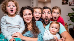 A smiling family of six sits close together on a couch, all with surprised, wide-eyed expressions and open mouths.