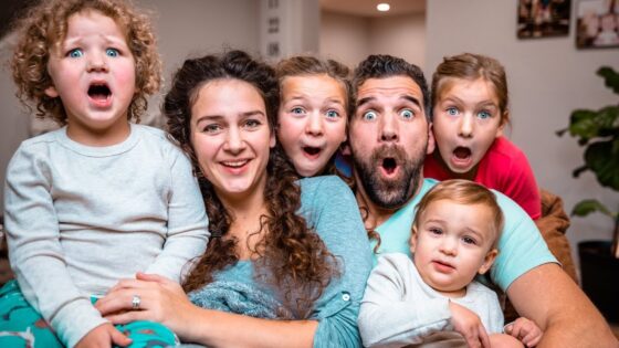 A smiling family of six sits close together on a couch, all with surprised, wide-eyed expressions and open mouths.