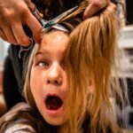 Young girl has her hair cut; a stylist uses scissors near her head with hair clipped up in preparation.