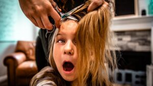 Young girl has her hair cut; a stylist uses scissors near her head with hair clipped up in preparation.
