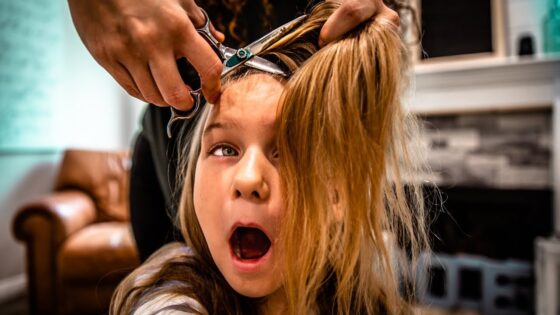Young girl has her hair cut; a stylist uses scissors near her head with hair clipped up in preparation.