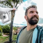 Bearded man in a green shirt takes a selfie as a white quadcopter drone hovers nearby in a suburban yard.