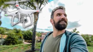 Bearded man in a green shirt takes a selfie as a white quadcopter drone hovers nearby in a suburban yard.