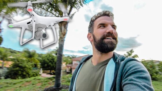 Bearded man in a green shirt takes a selfie as a white quadcopter drone hovers nearby in a suburban yard.