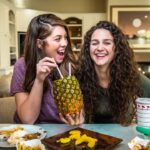 Two women sit at a dining table laughing as one sips from a pineapple with straws held between them in a bright kitchen/living room.